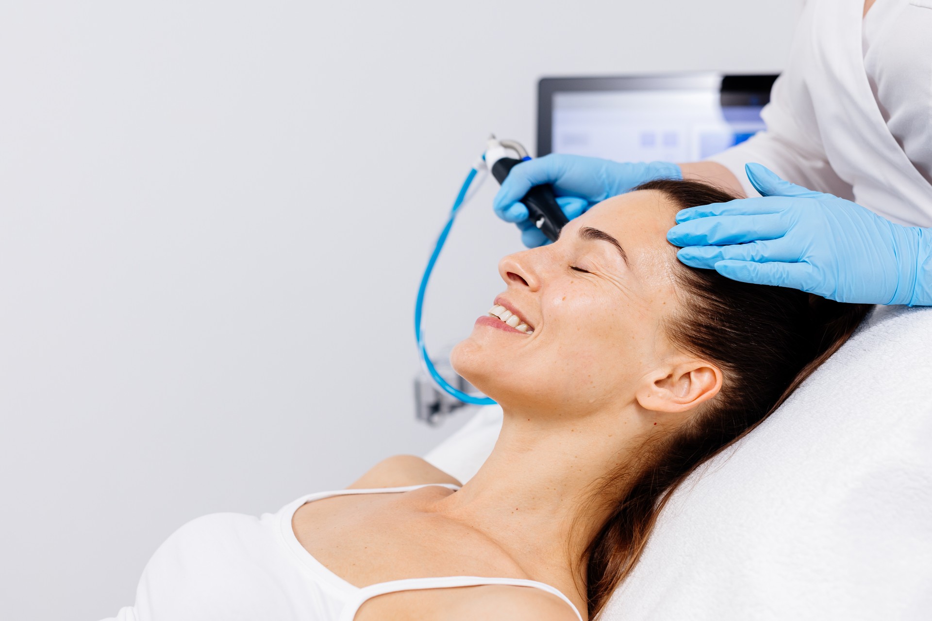 A middle-aged woman receives facial treatment in a professional beauty salon. A close-up cosmetologist manipulates a hydropeeling device to clean and rejuvenate facial skin.